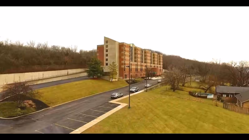 Exterior view of a multi-story senior living facility building with a parking lot and surrounding grassy areas, trees, and a few cars parked along the driveway. The building is situated near a wooded area.