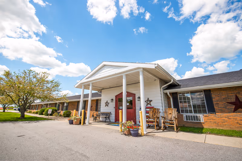 Exterior view of Signature HealthCARE of Trimble County building with a covered entrance supported by white columns, a red door, wooden chairs, potted plants, and a tree on the left side under a partly cloudy blue sky.