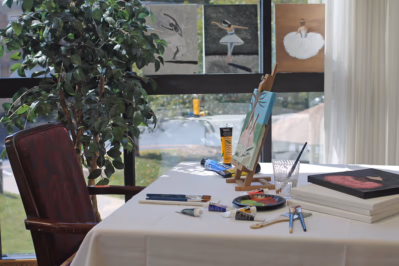 A bright art studio corner with a table covered in a white cloth, holding paintbrushes, tubes of paint, a glass of water with a brush, a small easel with a painting of pink rabbits, and several canvases. Behind the table is a large window with three paintings of ballerinas taped to it, and a leafy green plant next to a wooden chair with a red cushion.