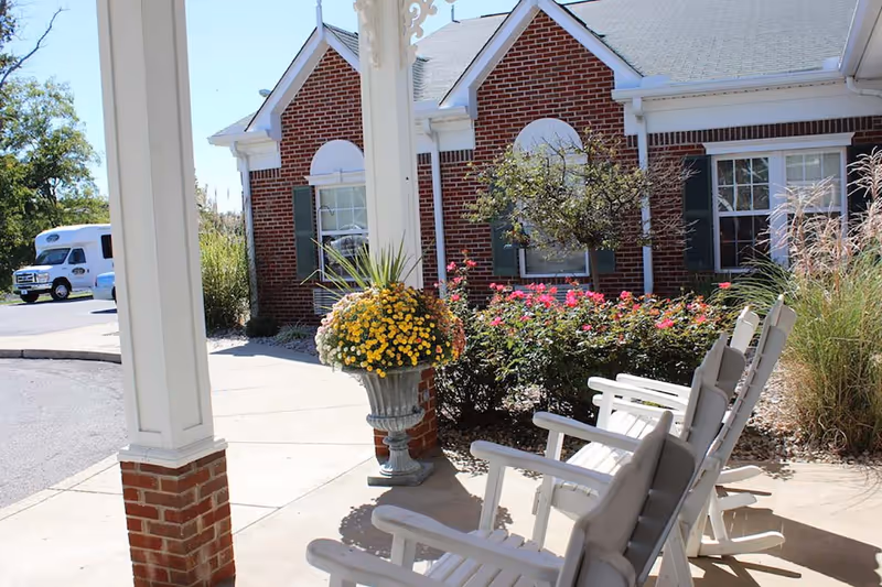 Outdoor seating area with white rocking chairs on a concrete patio next to a flower planter with yellow and white flowers, in front of a brick building with green shutters and windows. A small tree and pink flowers are visible in the garden bed nearby, and a white vehicle is parked in the background.
