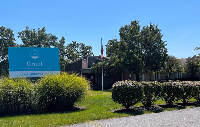 Exterior view of Grant Healthcare and Rehabilitation facility with a large blue sign displaying the facility name and address, surrounded by green bushes and trees under a clear blue sky.