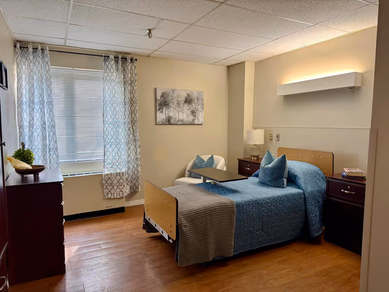 Senior living bedroom with single bed, blue quilt, nightstands, dresser, and window.