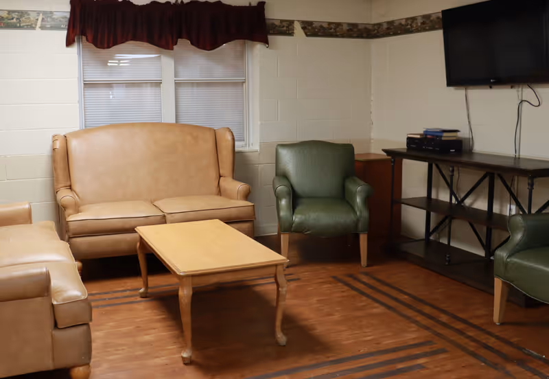 A cozy living room with a tan leather loveseat, two green armchairs, a wooden coffee table, a wooden floor, and a wall-mounted TV above a dark wooden console table. There is a window with closed blinds and a dark red valance above it.