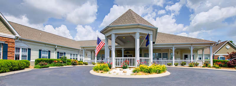 Front exterior view of The Bungalows at Mayfield facility featuring a covered entrance with white columns, American and state flags, surrounding landscaped flower beds, and a circular driveway under a partly cloudy sky.