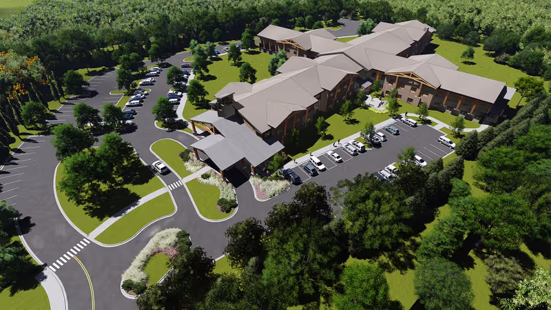 Aerial view of Boonespring Skilled Nursing Facility showing a large, multi-wing building surrounded by green lawns and trees. The facility has multiple parking lots with cars parked, and driveways with landscaped medians. The building has a light-colored roof and wooden architectural elements.