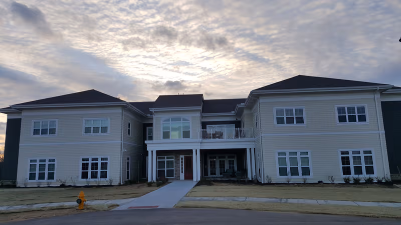 Two-story beige senior living facility with central entrance and balcony, under a cloudy sky.
