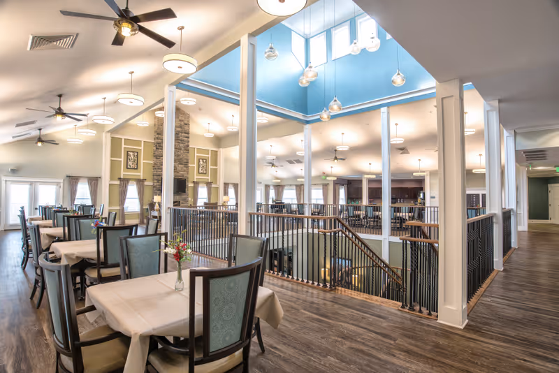 Bright, open two-level dining room with set tables and chairs, high ceilings, pendant lights, and a central staircase.