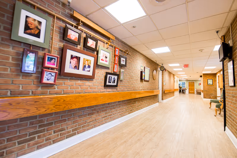 A well-lit hallway with brick walls adorned with framed photographs and wooden handrails. The floor is light-colored wood, and there are chairs along the right side. The hallway leads to a room with tables and chairs visible in the distance.