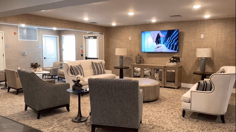 A cozy living room area in Creekside on Whipps Mill Road featuring a beige sofa with striped pillows, two gray armchairs, a round ottoman, side tables with lamps, a wooden cabinet with decorative items, and a wall-mounted TV displaying a close-up of hands opening a bottle.