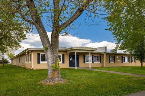 Single-story beige senior living facility with a covered entrance, lawn, and trees.