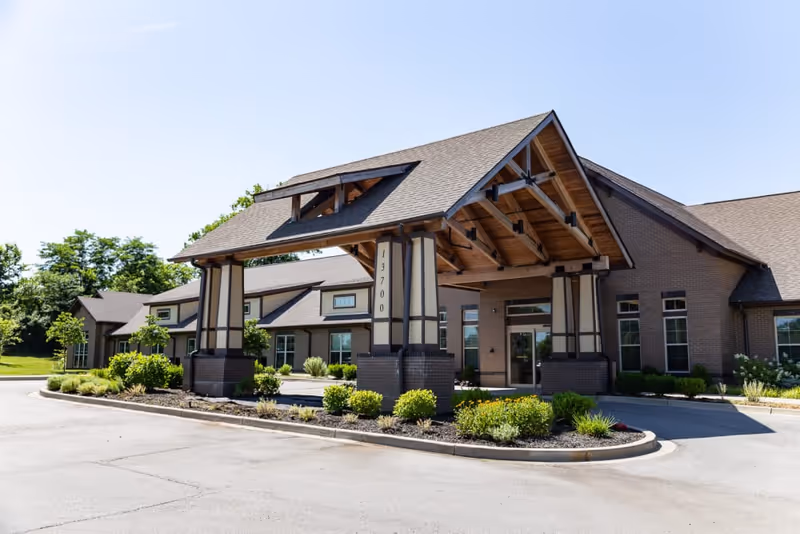 Exterior view of The Legacy at English Station building entrance with a covered driveway, landscaped plants, and clear blue sky.