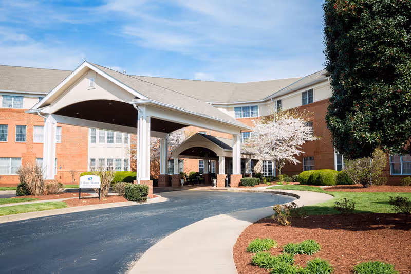 Exterior view of Village Manor senior living facility showing a large covered entrance with white pillars, a curved driveway, landscaped bushes, and blooming trees under a blue sky.