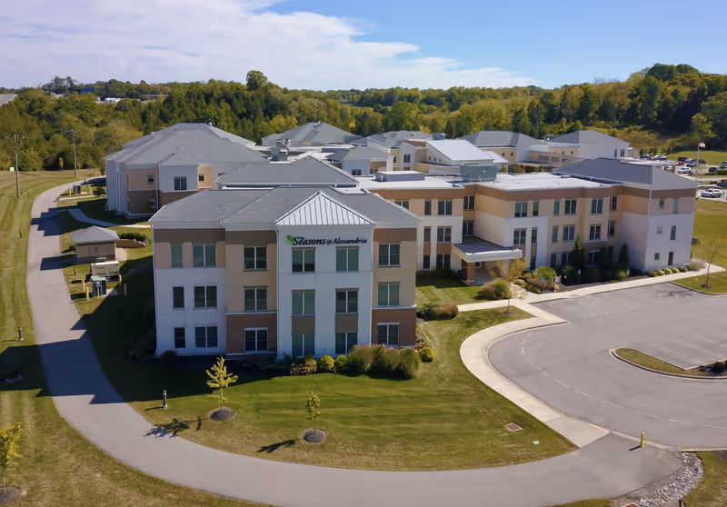 Aerial view of The Seasons@Alexandria senior living facility showing a large multi-story building with beige and white exterior walls, surrounded by green lawns, trees, and a curved driveway with parking spaces. The building is set against a backdrop of wooded hills under a partly cloudy sky.