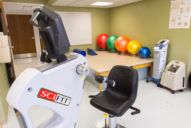 Rehabilitation room with a recumbent exercise bike, therapy table and colorful exercise balls against a green wall.