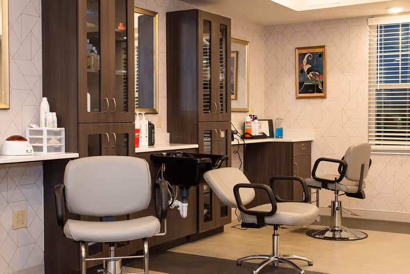 Interior view of a salon area in a senior living facility with three salon chairs, dark wood cabinets with glass doors, a black hair washing sink, various hair care products on the counters, a framed artwork on the wall, and a window with white blinds.