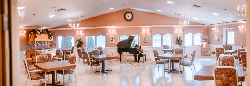 A bright senior living dining area with multiple tables, chairs, a grand piano, and windows.