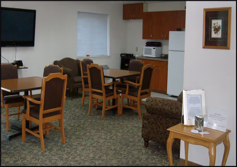 Communal room with dining tables and wooden chairs, a small kitchenette with a refrigerator and microwave, a wall-mounted TV, and upholstered seating.