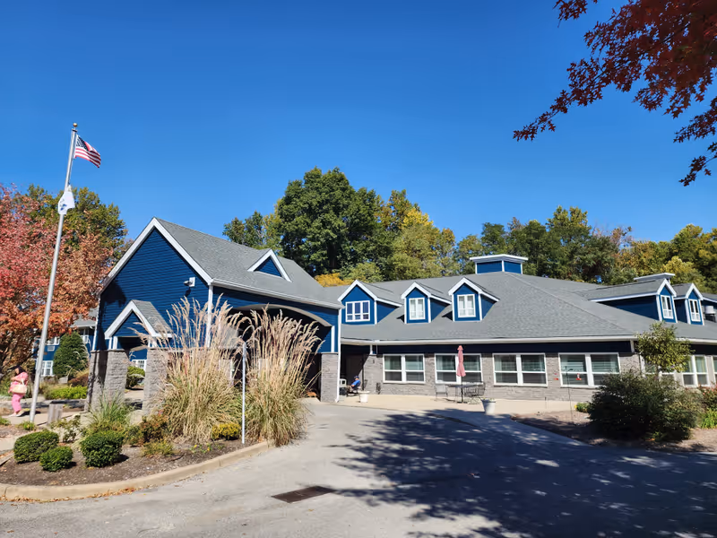 Exterior view of a senior living facility building with blue siding and gray roof under a clear blue sky. There is a covered entrance with stone pillars, an American flag on a flagpole, and landscaping with bushes and tall grasses. Trees with autumn foliage surround the building.