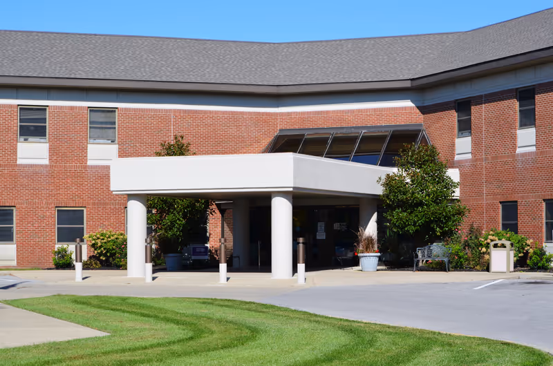 Front entrance of a two-story brick building with a covered drop-off area supported by white columns. There are windows on both floors, green shrubs and trees near the entrance, a bench, and a trash can. The sky is clear and blue.