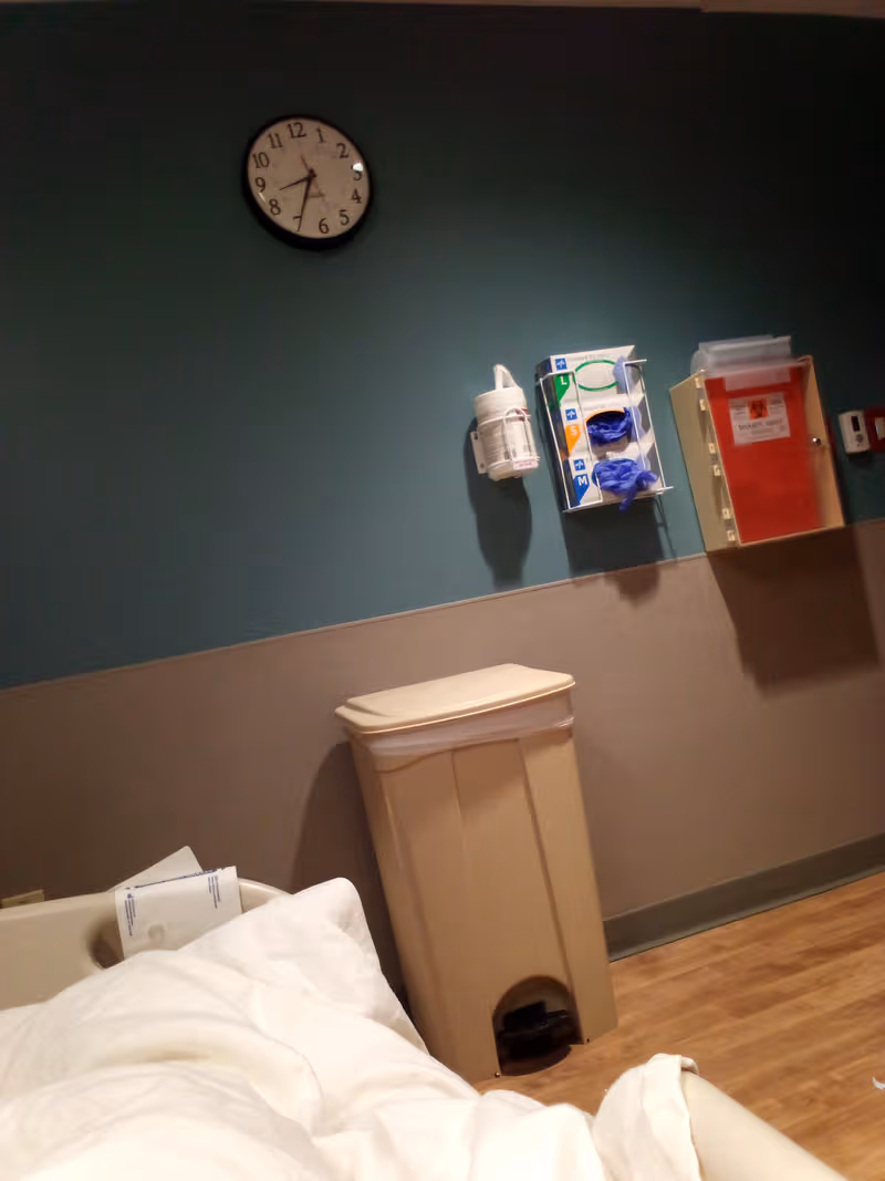Hospital-style patient room corner with bed, clock, wall-mounted supplies, and a trash can.