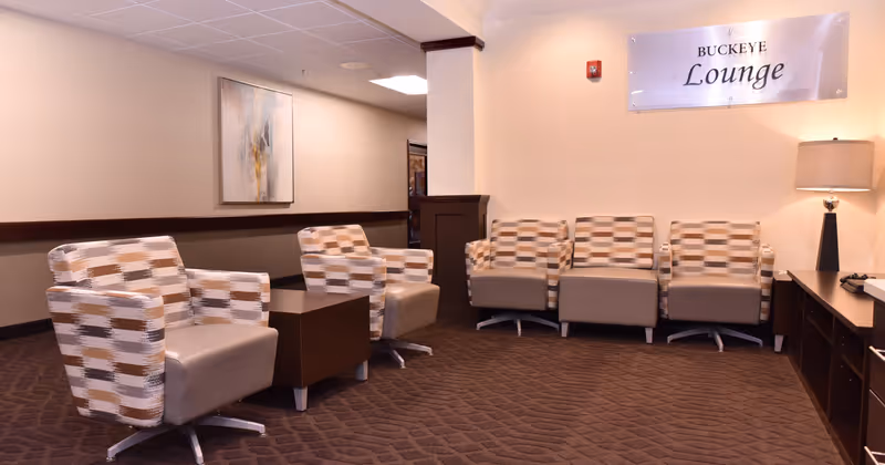A lounge area with five patterned armchairs arranged around a small wooden table. The room has beige walls, a brown carpet, a lamp on a wooden cabinet, and a sign on the wall that reads 'BUCKEYE Lounge'. There is also a framed abstract painting on one wall.