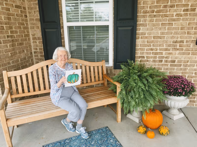 An elderly woman with white hair and glasses sits on a wooden bench outside a brick building. She is holding a drawing of a green and orange pumpkin. Next to the bench are a large green fern, a pot of purple flowers, and several pumpkins arranged on the ground. A blue patterned rug is on the concrete floor in front of the bench.