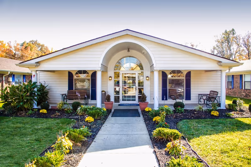 Front exterior view of a single-story building with white siding and a central entrance featuring a glass door under an arched window. There are two benches on either side of the entrance, surrounded by landscaped flower beds with green plants and yellow flowers. The building is part of a senior living facility with a well-maintained lawn and neighboring buildings visible.