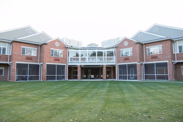Two-story red-brick senior living building with screened lower patios, a central covered walkway and upper balcony overlooking a large lawn.