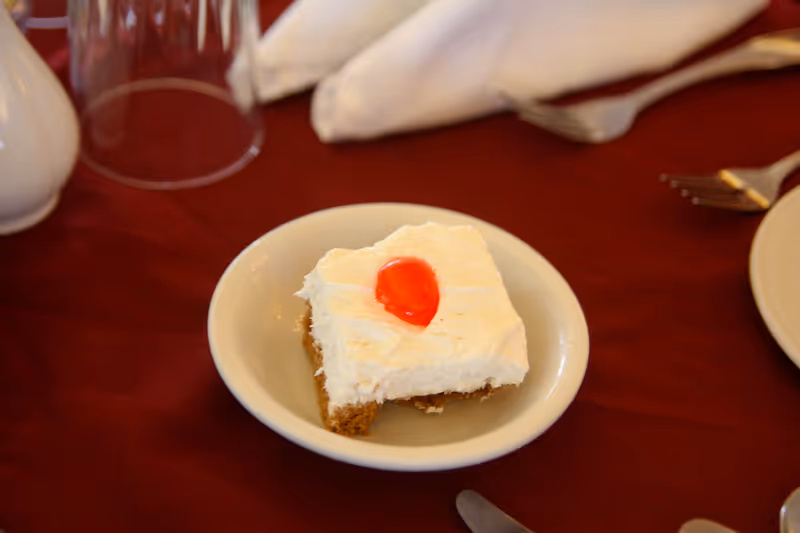 A small square piece of dessert with white frosting and a red cherry on top, served on a white plate placed on a red tablecloth. Nearby are a folded white napkin, a fork, a knife, and an upside-down clear plastic cup.