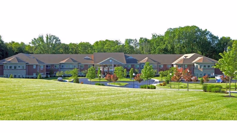 Front exterior view of Magnolia Springs East Louisville, a large two-story senior living facility surrounded by green grass, trees, and a circular driveway with parked cars.