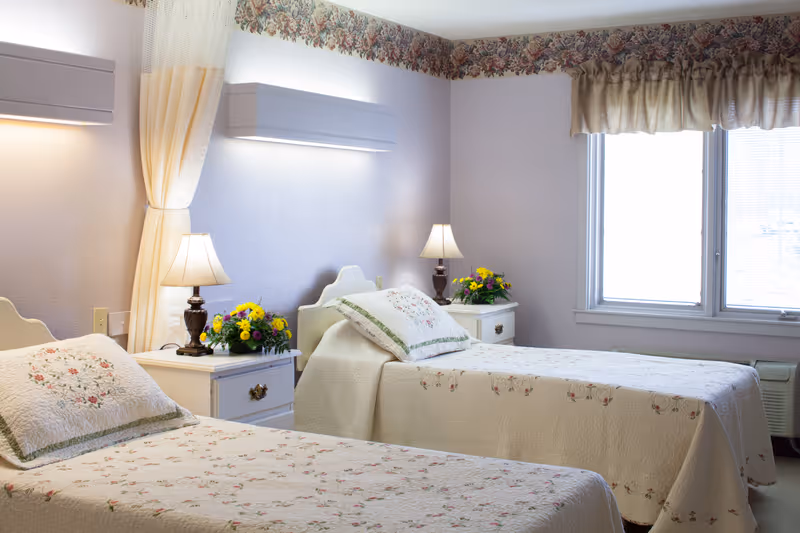 A bright and cozy bedroom with two twin beds covered in cream-colored floral quilts. Each bed has a matching pillow and a white nightstand beside it with a lamp and a small bouquet of flowers. The walls are light purple with floral wallpaper border near the ceiling, and a window with beige curtains lets in natural light.