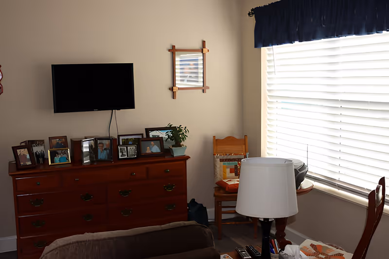 A cozy living room corner with a wooden dresser topped with multiple framed photos and a small potted plant. A flat-screen TV is mounted on the wall above the dresser. Next to the dresser is a wooden chair with a colorful quilted cushion, positioned near a window with white blinds and a dark valance. In the foreground, there is a table lamp and part of a brown sofa.