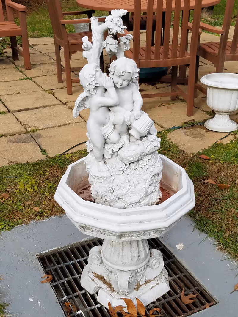 White cherub statue fountain on a paved outdoor patio with wooden chairs.