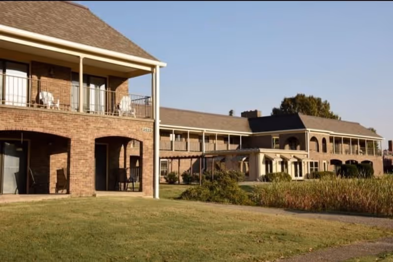 Exterior view of a two-story brick assisted living apartment building with balconies and patios, surrounded by a grassy lawn and some bushes under a clear sky.