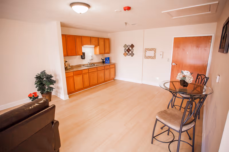 Open studio living area with a kitchenette along the back wall, a glass-top dining table with chairs, and wood flooring.