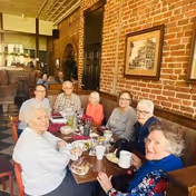A group of seven elderly people sitting around a wooden table in a restaurant with exposed brick walls, enjoying a meal and drinks together. The setting includes framed pictures on the wall and a warm, cozy atmosphere.