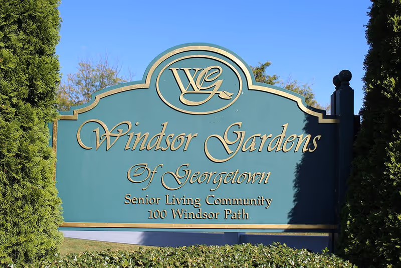 A green and gold sign for Windsor Gardens of Georgetown Senior Living Community located at 100 Windsor Path, surrounded by greenery and trees under a clear blue sky.