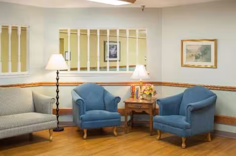 Cozy senior living lounge with blue chairs, a sofa, lamps, and a small table.