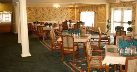 Dining room with multiple wooden tables and chairs arranged neatly. Each table is set with folded green napkins and glassware. The room has floral wallpaper, carpeted floors with a patterned rug, and large windows with curtains allowing natural light to enter. There is a double door exit visible in the background.