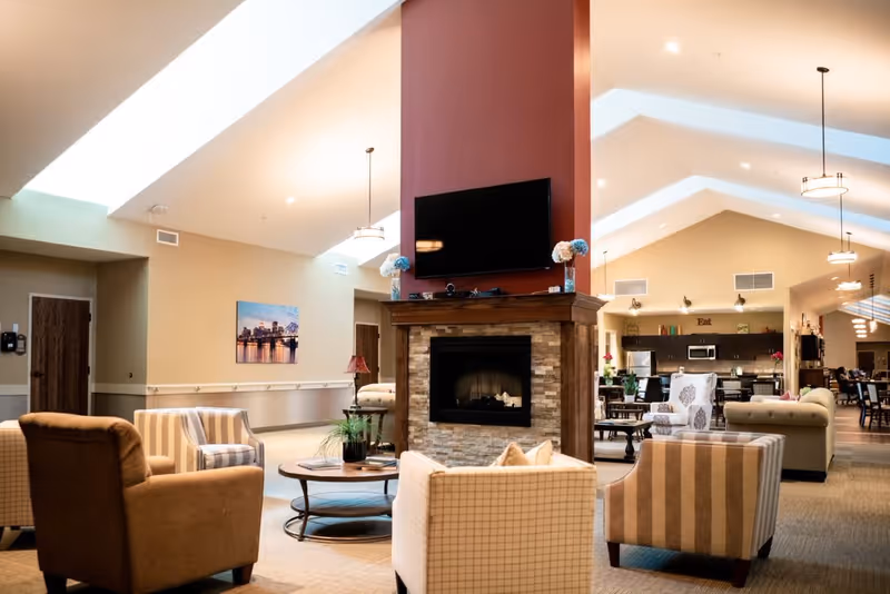 A spacious senior living facility common area with a central stone fireplace topped by a mounted flat-screen TV. Surrounding the fireplace are various comfortable armchairs and a round coffee table with plants and books. The room has high vaulted ceilings with skylights and pendant lights. In the background, there is a kitchen area with stainless steel appliances and a dining area with tables and chairs.