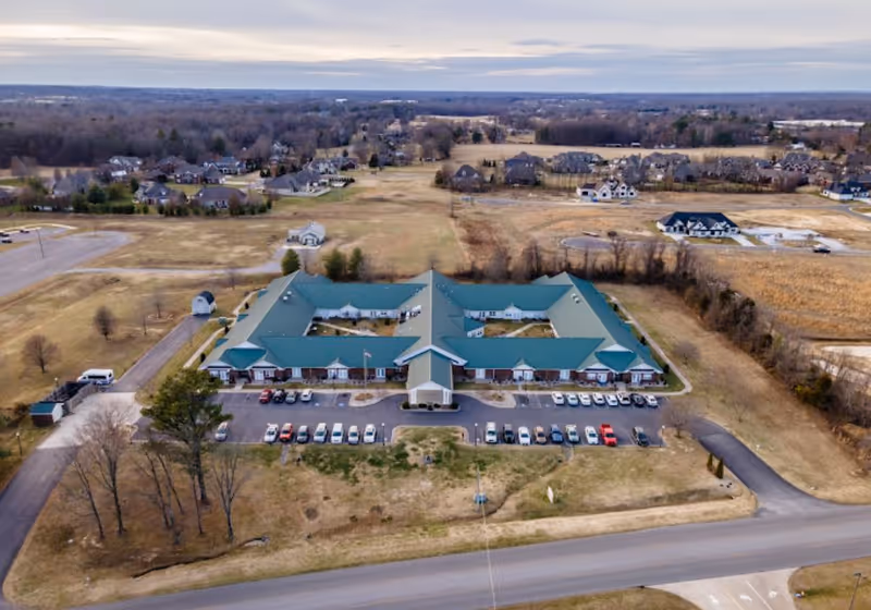 Aerial view of a single-story senior living facility with green roofs, a central entrance and parking lot surrounded by open fields and nearby houses.