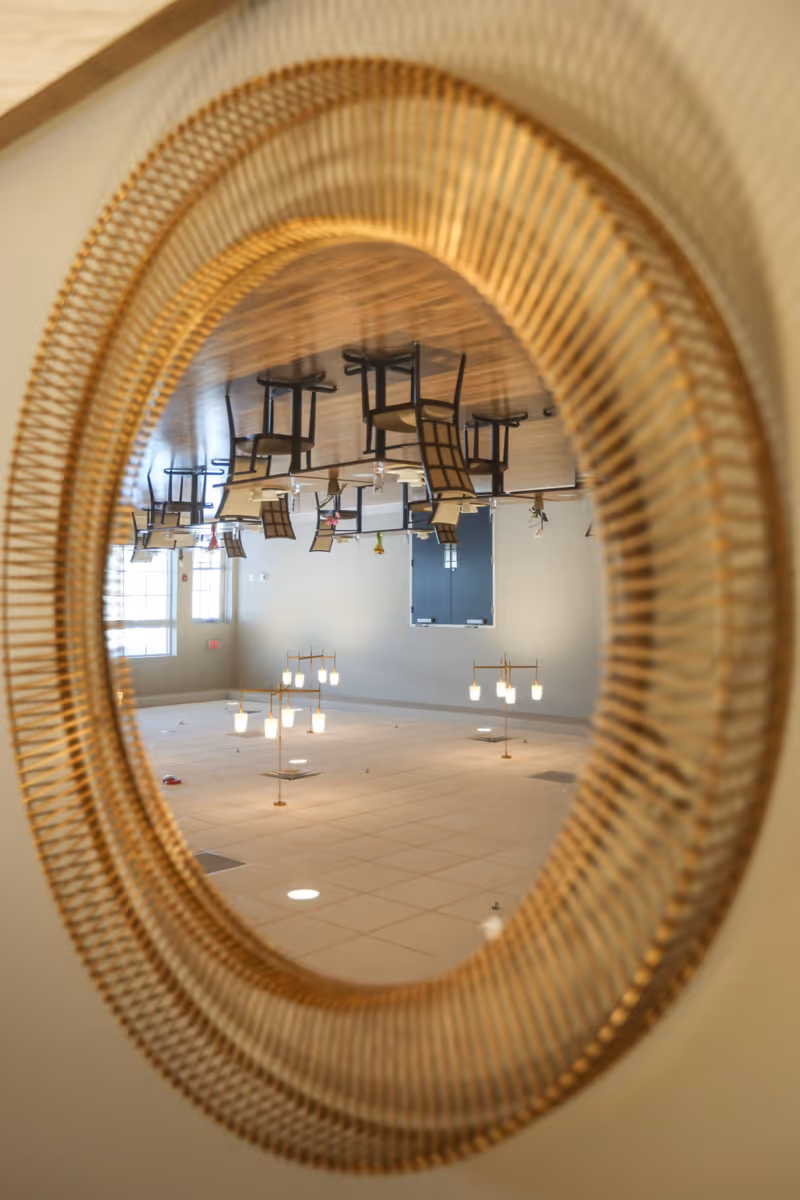 Reflection in a round, gold-framed mirror showing an empty dining area with wooden floor, tables, and chairs. The ceiling with lights and vents is also visible in the reflection.