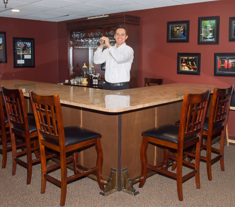 A man in a white shirt stands behind a large marble-topped bar counter shaking a cocktail shaker. The bar has several wooden chairs with black cushioned seats around it. Behind the man is a wooden cabinet with glassware and bottles. The walls are painted dark red and decorated with framed pictures.