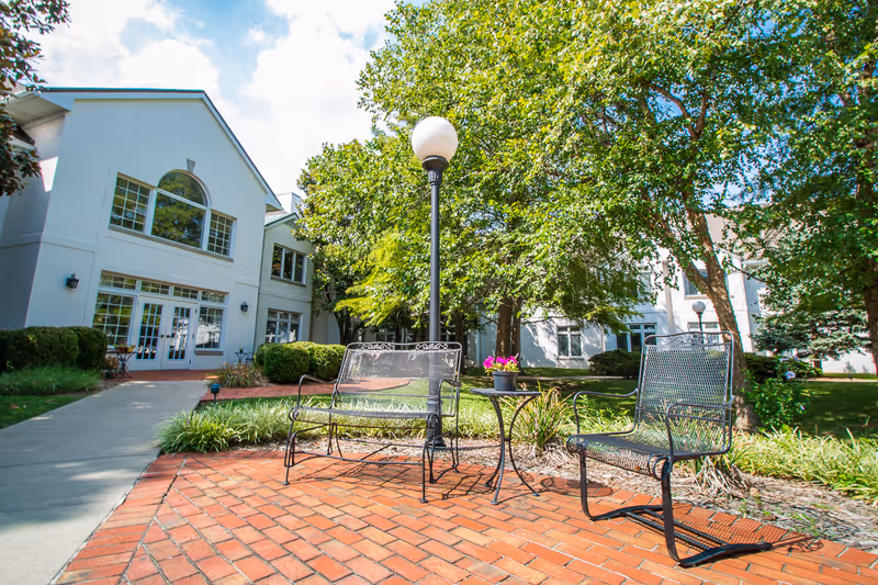 Outdoor seating area with two metal chairs and a small table with a potted plant on a brick patio. The area is surrounded by greenery and trees, with a white building in the background under a partly cloudy sky.