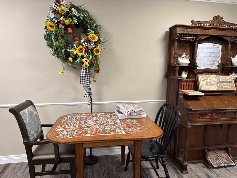 A wooden table with two chairs, one upholstered and one black wooden, with a partially completed jigsaw puzzle and puzzle box on top. Behind the table is a decorative floral wreath with sunflowers and red flowers hanging on a beige wall. To the right is an ornate wooden cabinet with vintage sheet music and ceramic teapots displayed.