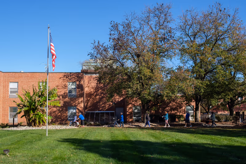 Front exterior of a brick building with an American flag, large trees, and several people walking along a path across a grassy lawn.