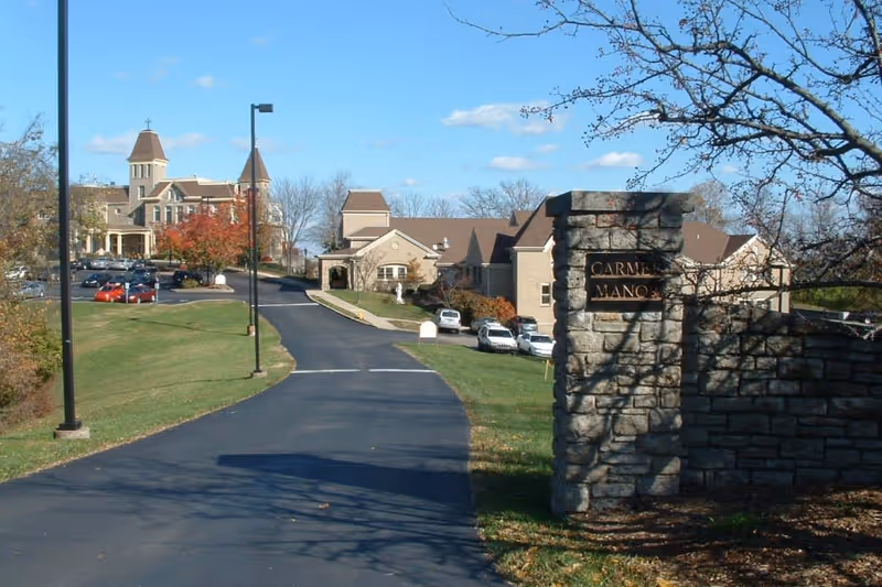 View of Carmel Manor facility showing a paved driveway leading up to multiple buildings with a parking lot on the left. The buildings have a traditional architectural style with towers and pitched roofs. A stone sign with the text 'Carmel Manor' is visible on the right side near the entrance.