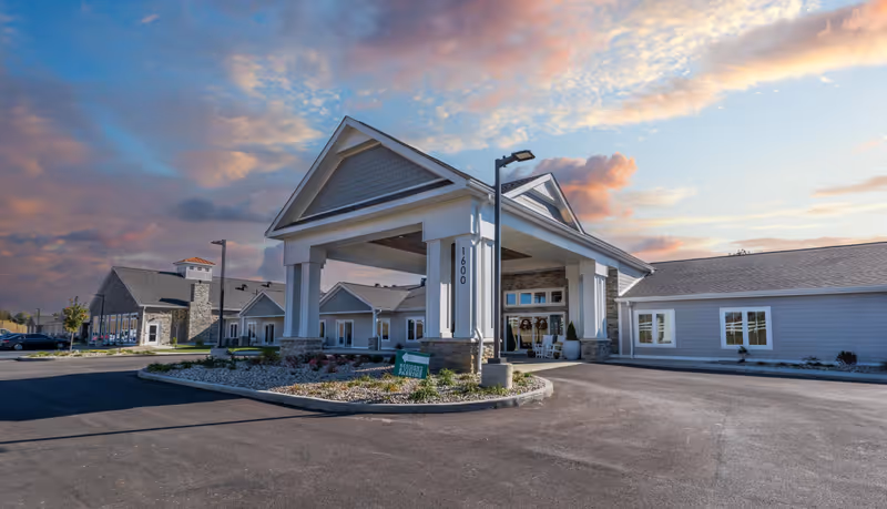 Exterior view of Cedarhurst Senior Living of Nicholasville building at sunset, showing the main entrance with a covered drop-off area, landscaped surroundings, and a clear sky with scattered clouds.