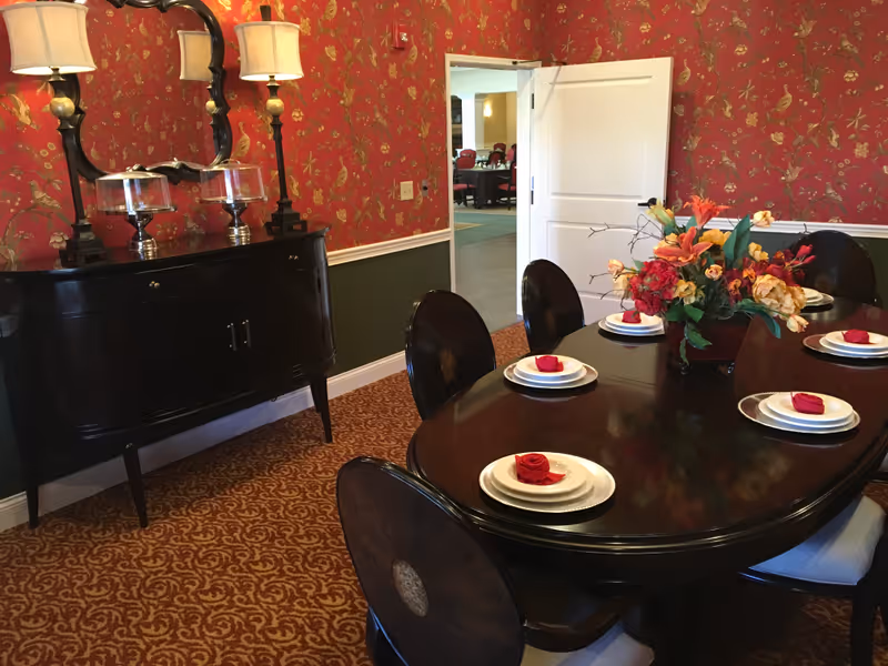 A formal dining room with a dark wooden oval table set with white plates and red napkins shaped like roses. A floral centerpiece decorates the middle of the table. The room has red wallpaper with a gold bird and floral pattern, a patterned carpet, and a black sideboard with two lamps and a mirror above it. An open door leads to another room with tables and chairs.