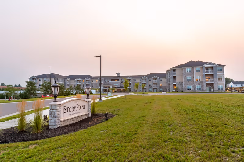 Exterior view of the StoryPoint Union senior living facility at sunset, showing a large multi-story building with balconies, a well-maintained lawn, a paved driveway, and a stone sign with lamps displaying the facility name.